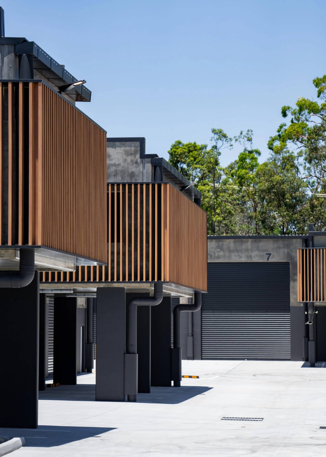 Modern commercial factory units with timber screens and dark roller doors opening onto a landscaped concrete hardstand under a clear sky.