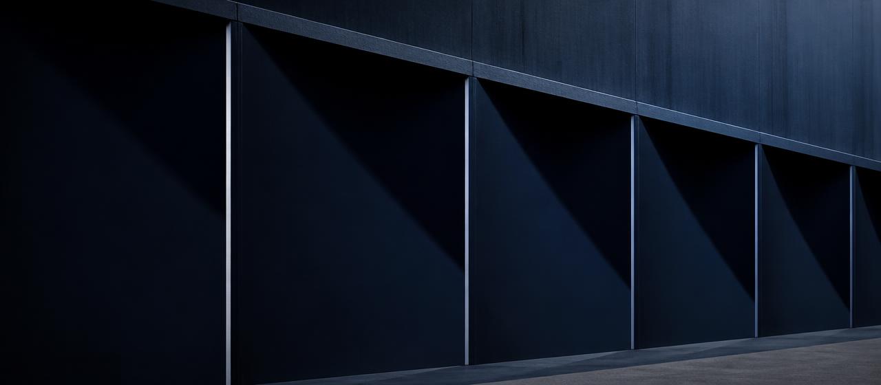 Dark steel facade in repeating structural bays, each edge throwing a sharp diagonal shadow.