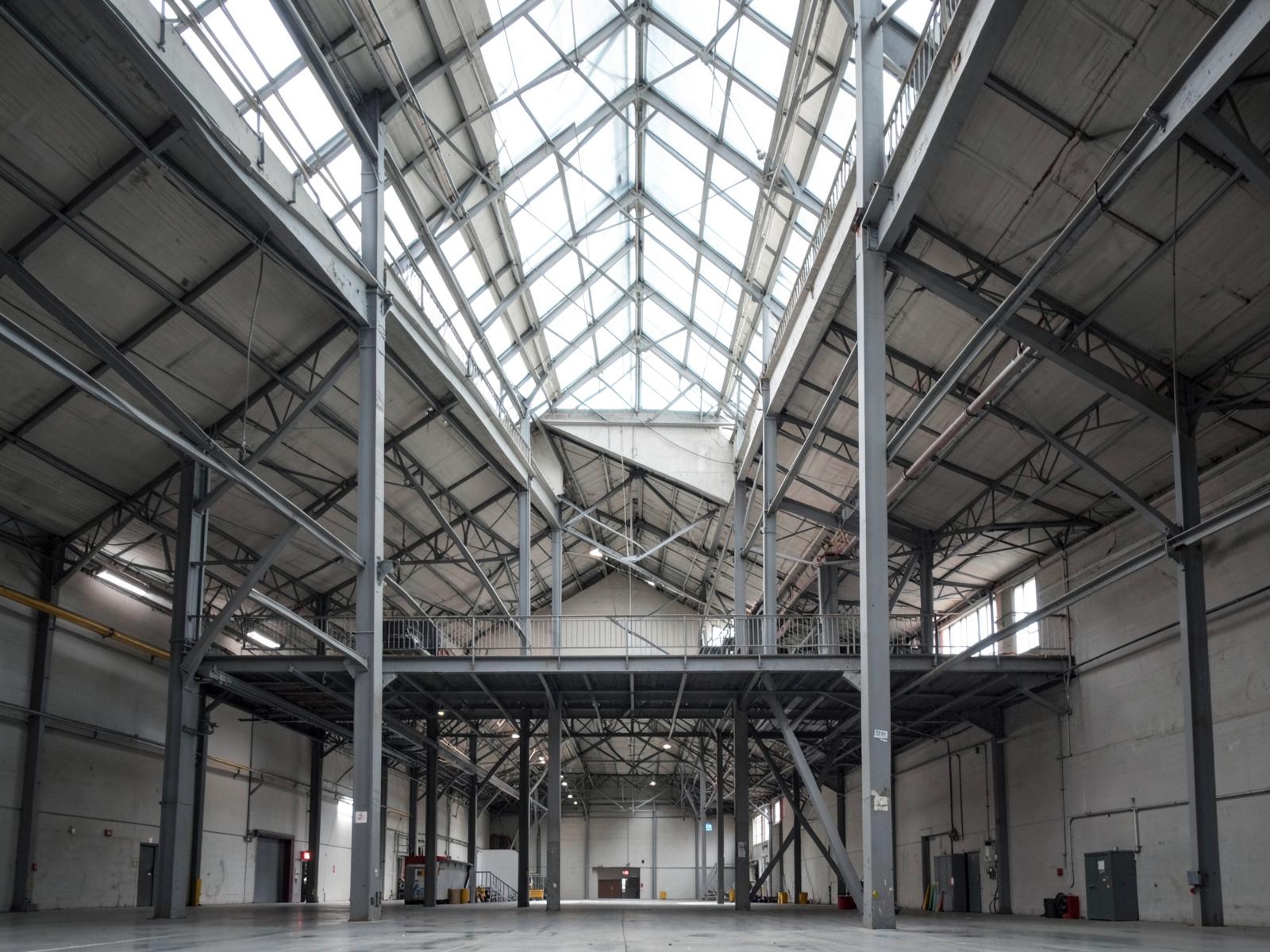 Tall clear span industrial interior with exposed portal frames and a mezzanine.